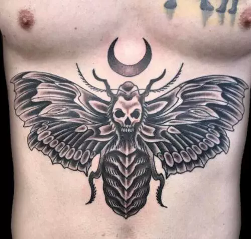 A detailed black and gray tattoo of a moth with a skull face and a crescent moon above it, inked on a person's chest.