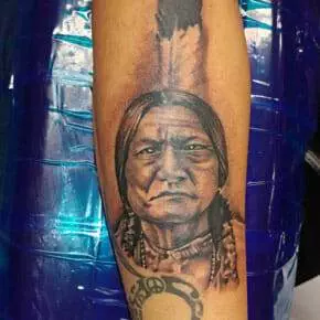 Forearm tattoo of a Native American man with long hair, wearing traditional attire, in black and grey ink. A feather is tattooed above the man's head.