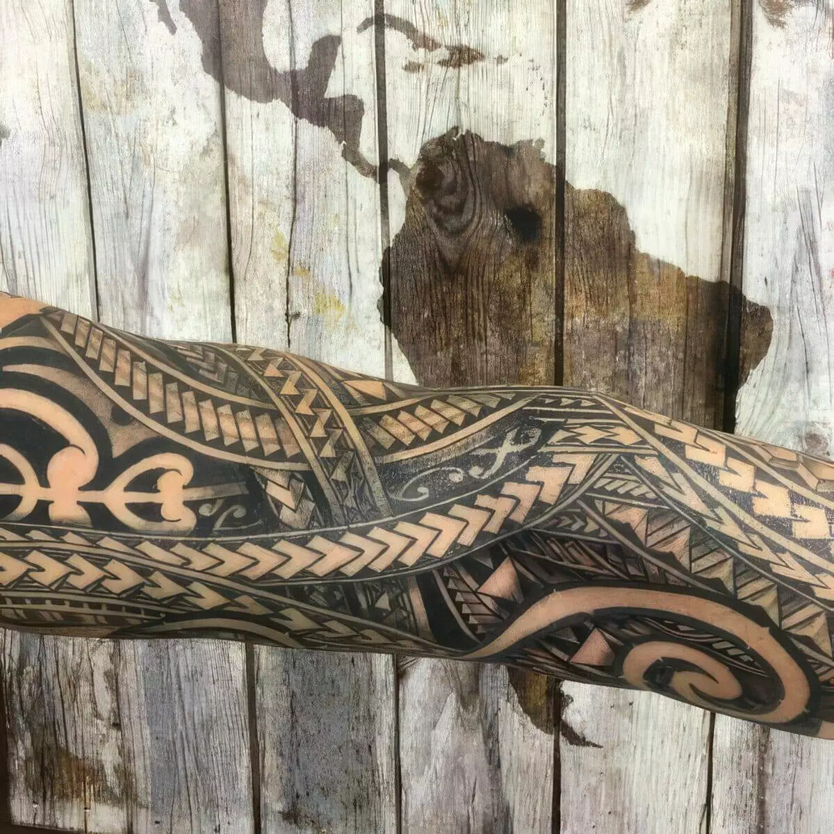 An intricately designed tattoo covers a person's forearm, featuring geometric and tribal patterns in black ink, with a faded wooden background depicting a world map.