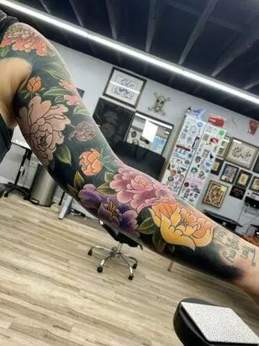 A colorful floral tattoo sleeve on a person's arm, depicted in a tattoo studio with various artworks on the walls and a wooden floor.