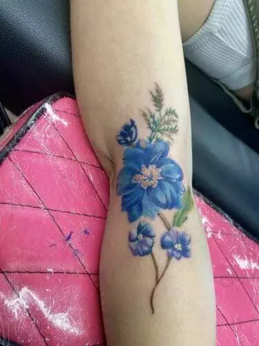 A colorful tattoo featuring blue flowers and green leaves is displayed on a person's forearm.