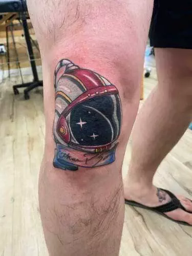 A tattoo of an astronaut's helmet with a nebula visible inside, located on a person's knee.