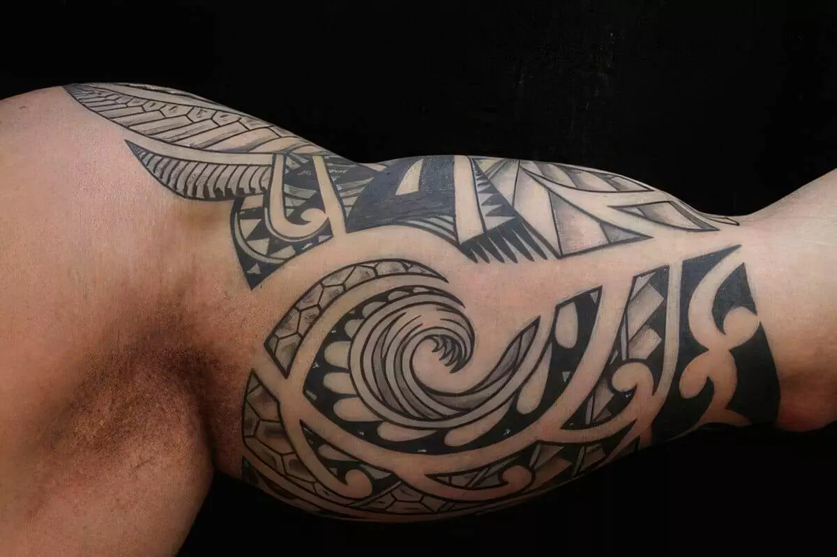 A close-up of a flexed arm with an elaborate black tribal tattoo covering the bicep, featuring geometric patterns and spiral designs typical of Polynesian art.