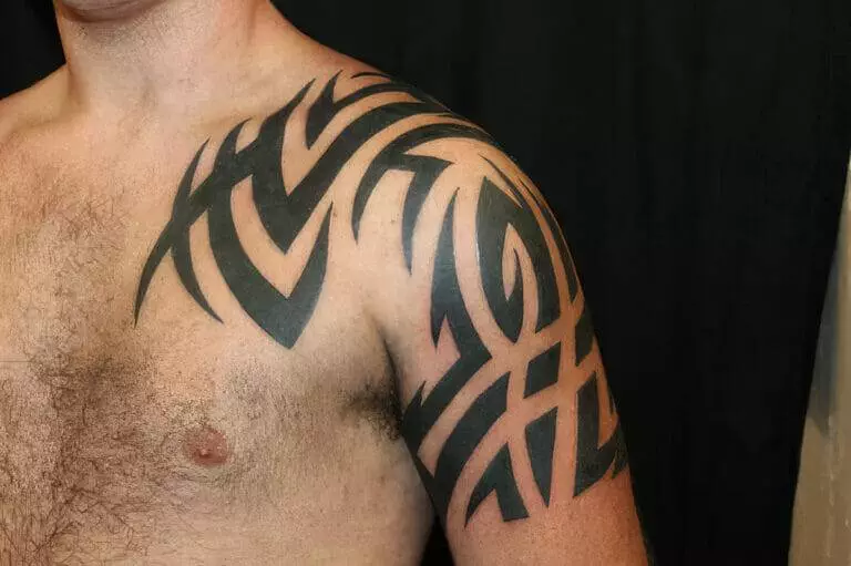 A close-up of a man's upper body showing a black tribal tattoo covering his shoulder, upper arm, and part of his chest.