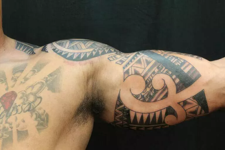 A close-up of a person's arm extended sideways, showcasing an intricate tribal tattoo design on the bicep and a partially visible chest tattoo.