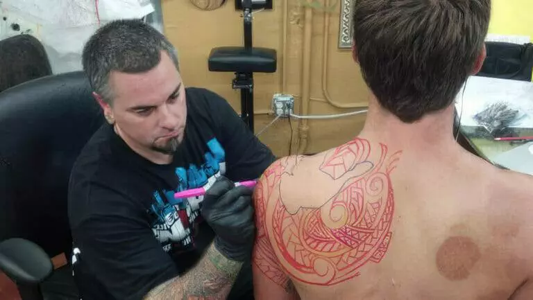 A tattoo artist is inking a large tribal design in red on the back and shoulder of a shirtless person sitting in a chair.