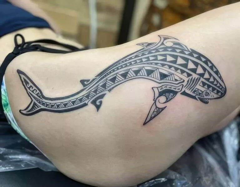 A person with a large, intricate shark tattoo featuring geometric patterns on their lower back and hip. The person is lying down during the tattooing process.