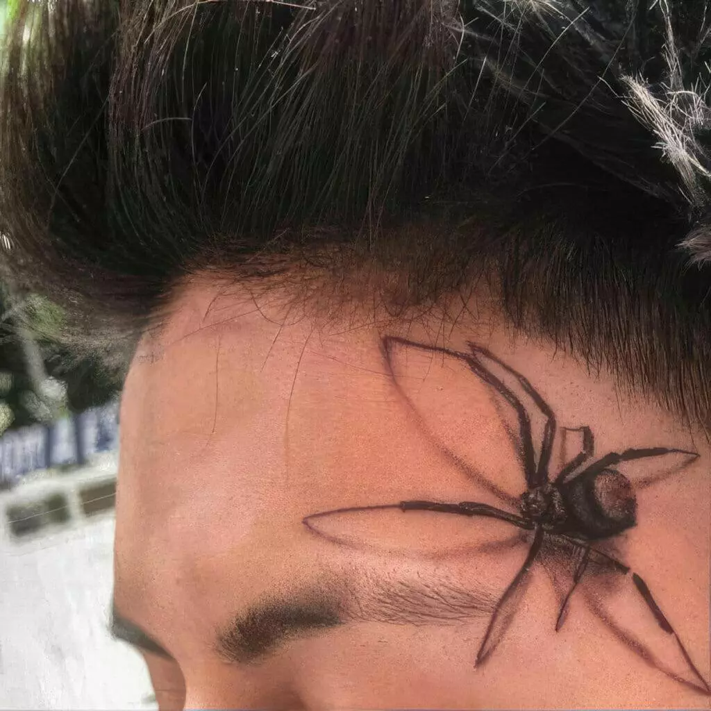 A close-up of a person's forehead with a large, realistic black spider tattoo near the temple and eyebrow.