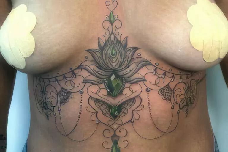 A detailed, symmetrical chest tattoo features ornamental designs, floral motifs, and green gem-like elements. Beige tape strips are visible on both sides of the chest.