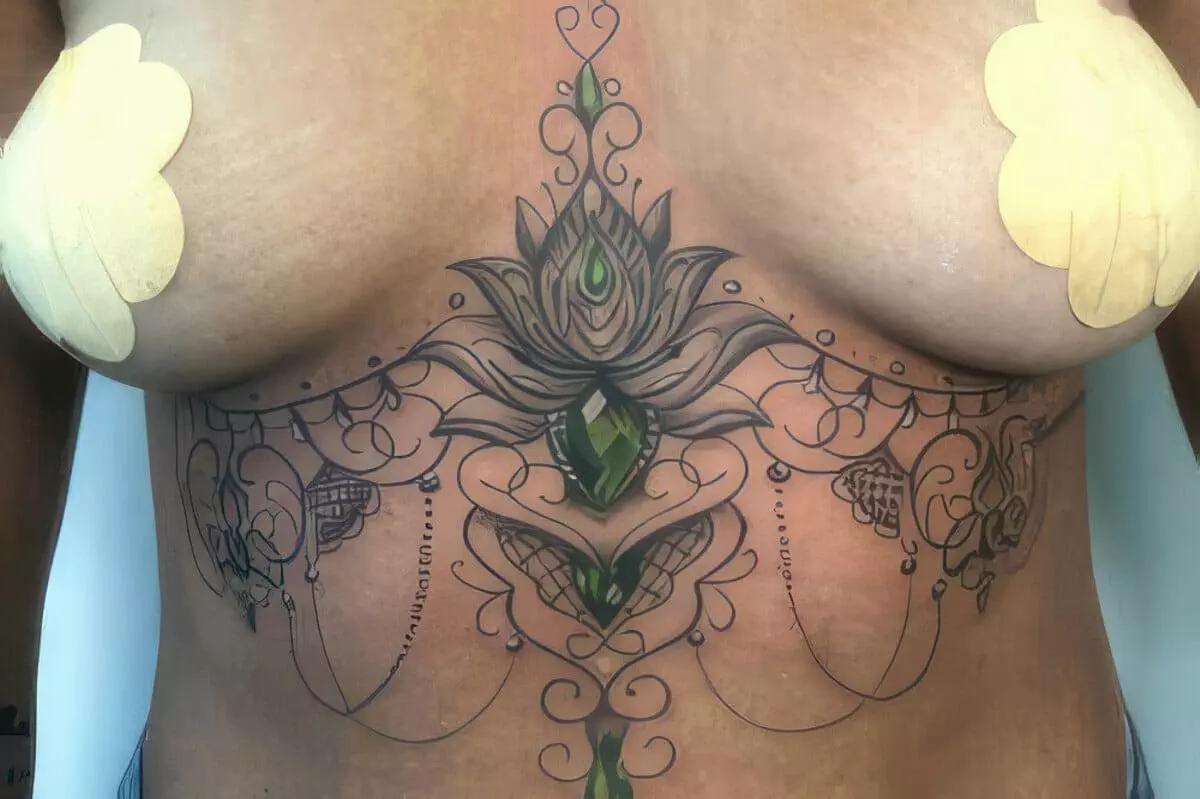 A detailed, symmetrical chest tattoo features ornamental designs, floral motifs, and green gem-like elements. Beige tape strips are visible on both sides of the chest.