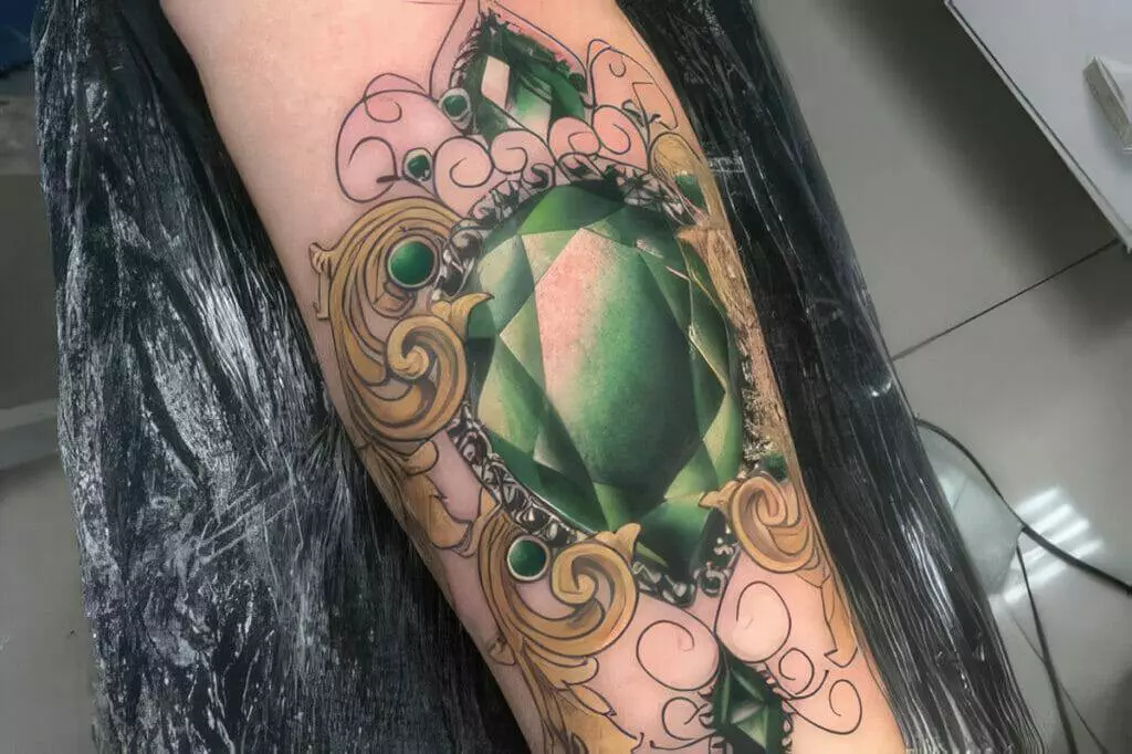 A detailed tattoo of a large green gemstone with ornate designs surrounding it, located on a person's thigh.