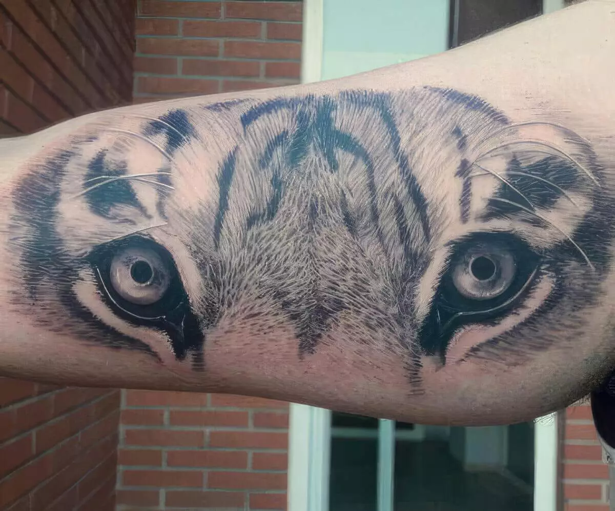 A tattoo of a realistic tiger's eyes and forehead is inked on someone's upper arm, with a brick wall in the background.