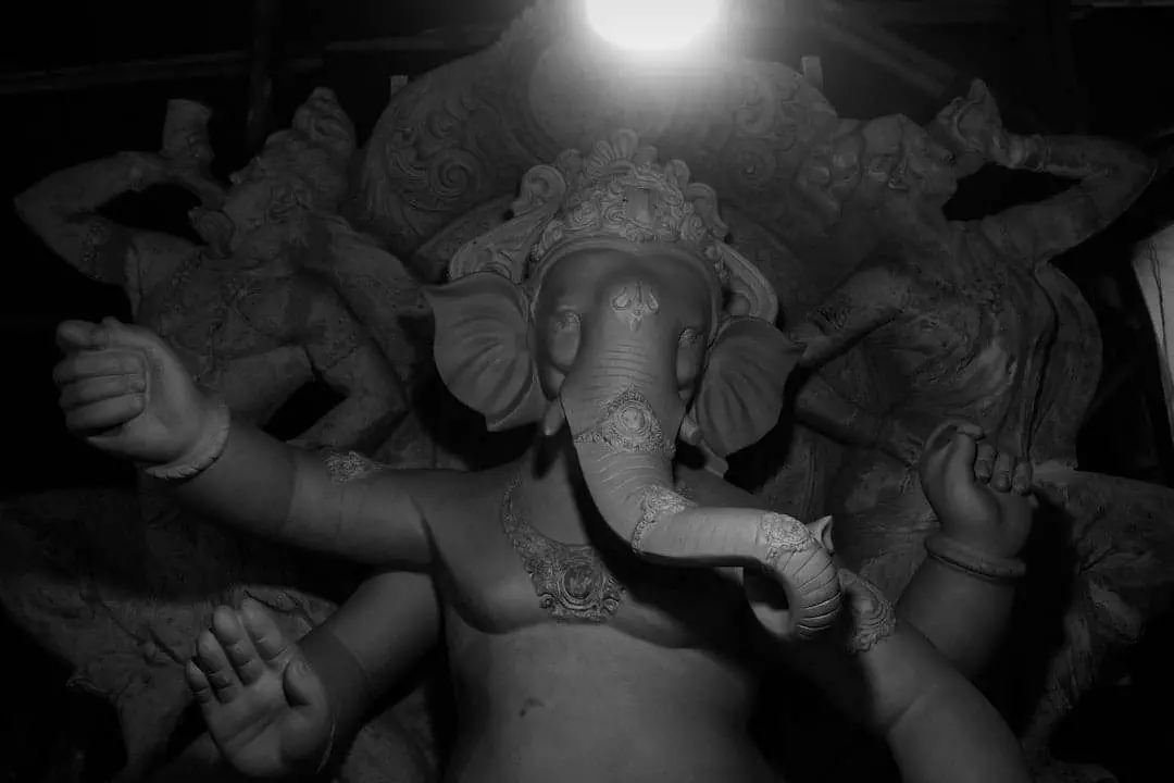 Black and white image of a statue of Ganesha with detailed carvings. Two additional figures are positioned behind the main statue. The image is illuminated from above.