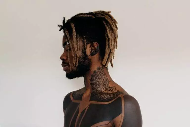 Profile view of an individual with blond-tipped dreadlocks and intricate black tattoos covering their neck and shoulders, standing against a plain white background.