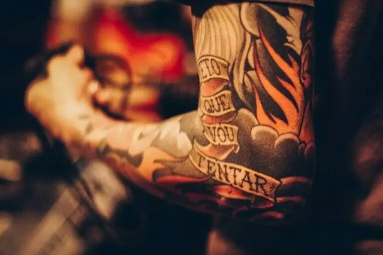 A close-up of a person's arm covered with colorful tattoos, including text on a banner that reads, 