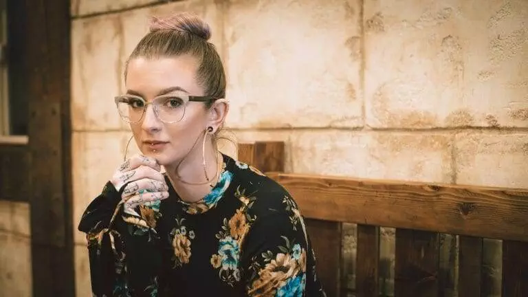 A person with glasses, wearing a floral top, sits on a wooden bench against a textured wall, resting their chin on their hand and looking at the camera.