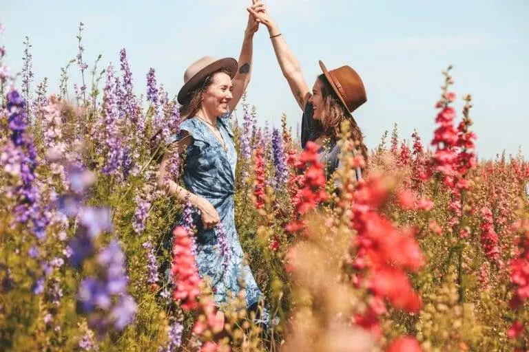 Two people in hats are holding hands and smiling in a field of colorful flowers, including red, purple, and pink blooms, on a sunny day.