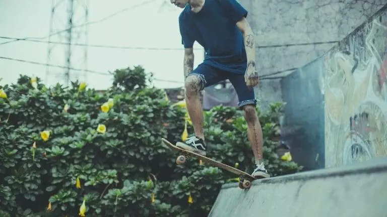 Person in blue shirt and denim shorts skateboarding on a ramp near graffiti-covered wall and greenery with yellow flowers.