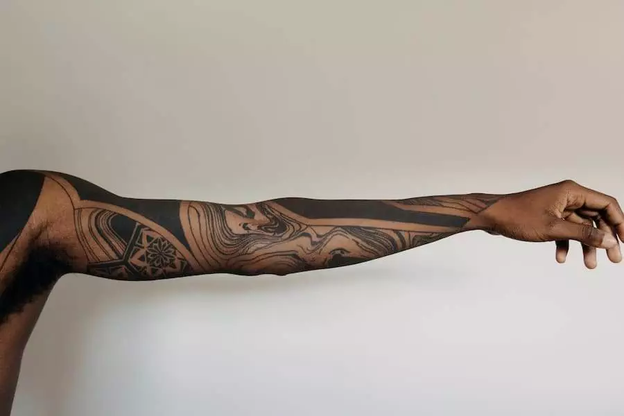 A person's extended arm showing black ink tattoos with abstract designs against a plain background.