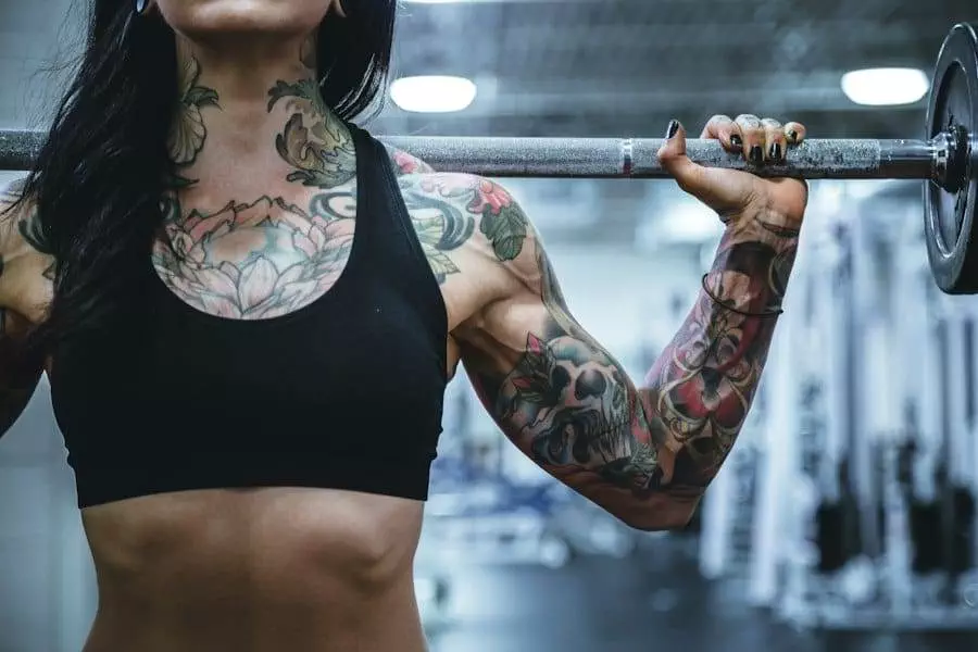 A person with extensive arm and chest tattoos holds a weighted barbell across their shoulders in a gym.