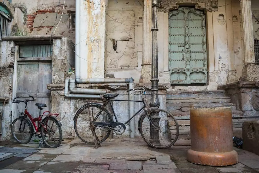 Two old bicycles are parked against a weathered building, featuring an ornate green door, and peeling paint on its walls. A large rusty metal cylinder sits nearby on the stone-paved ground.