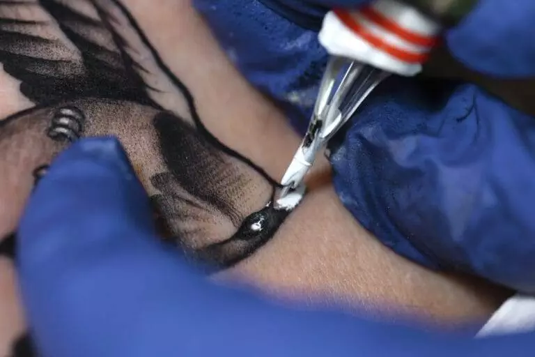 Close-up of a tattoo being applied to the skin by a tattoo artist wearing blue gloves. The tattoo design appears to be a bird with visible black ink details.