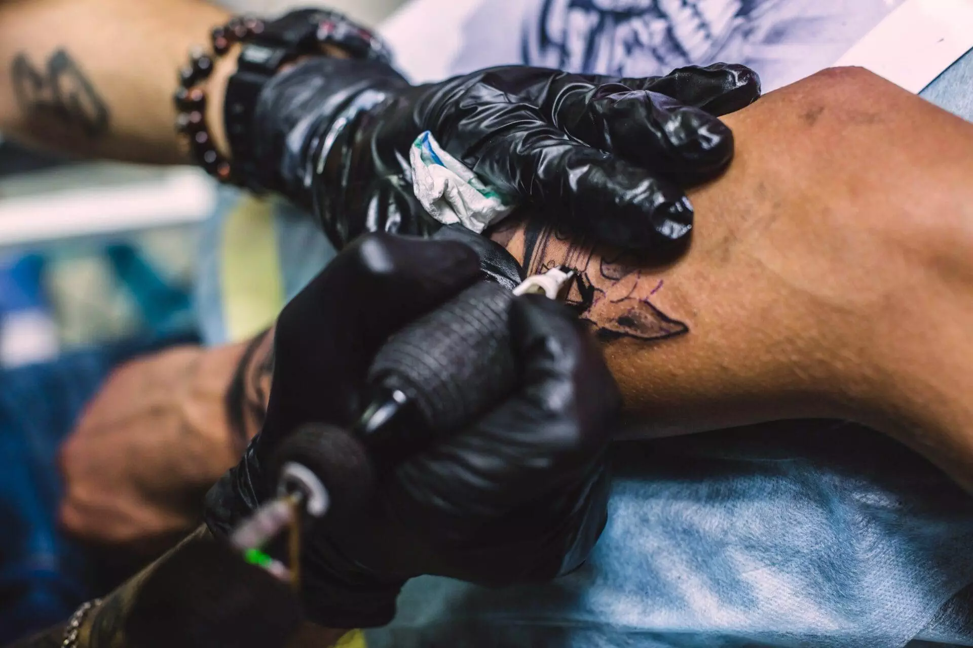 A tattoo artist wearing black gloves creates a tattoo design on a person's forearm using a tattoo machine.