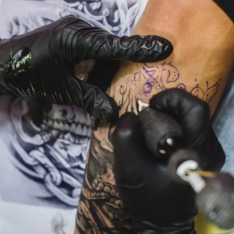 A tattoo artist wearing black gloves works on a tattoo design on a person's arm, using a tattoo machine.