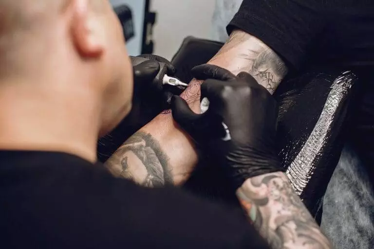 An artist with black gloves is tattooing a person's forearm. The tattoo equipment and the person's existing tattoos are visible.