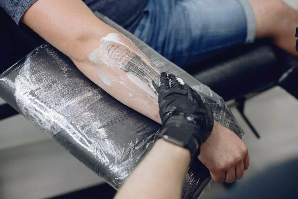 A person’s forearm is being prepped for a tattoo with cream. The person is seated, and a gloved hand is applying the cream while holding the arm steady.