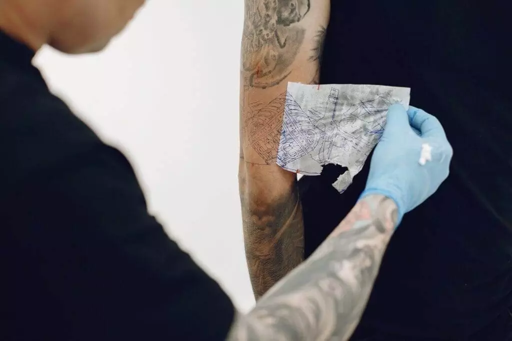 A tattoo artist wearing gloves places a stencil on a person's arm, preparing for a tattoo design.