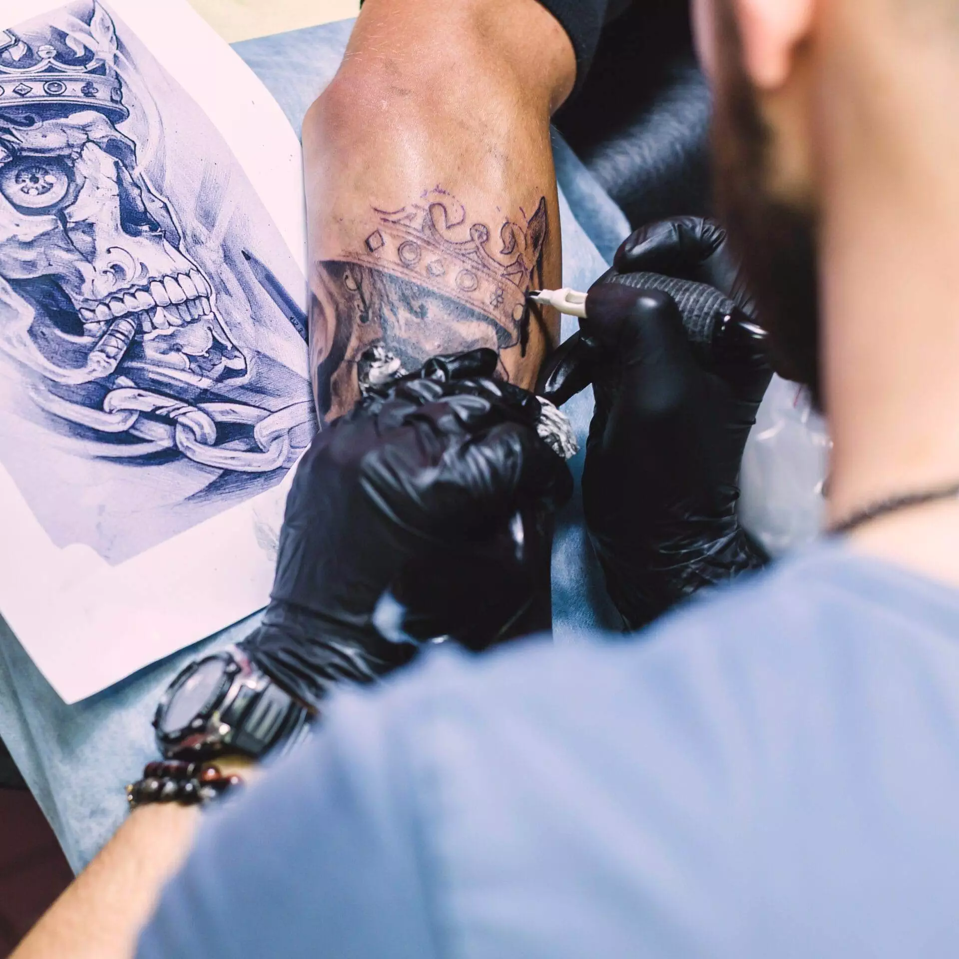 A tattoo artist with black gloves tattoos a detailed skull and chain design on a person's forearm, using a reference image nearby.