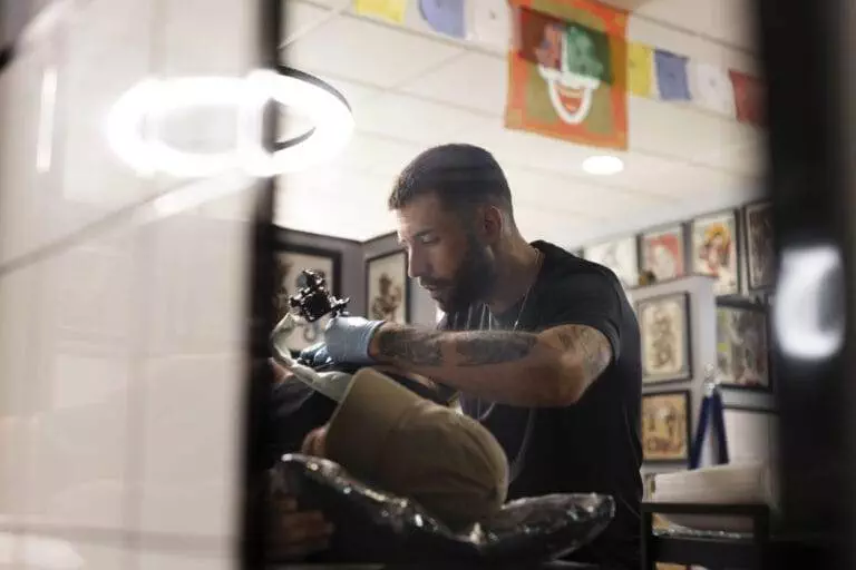 A tattoo artist with a beard is focused on tattooing a person's arm in a studio decorated with various artworks and flags.