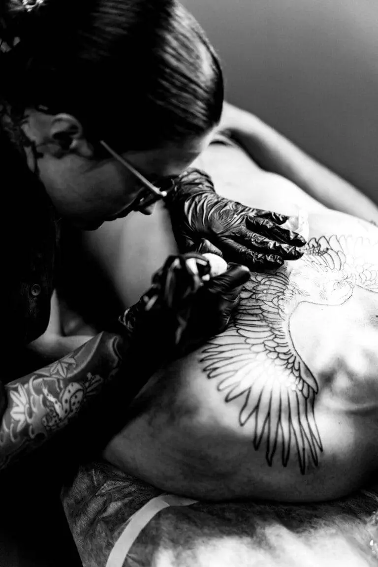 A tattoo artist wearing gloves is tattooing an intricate bird design on a person's back.