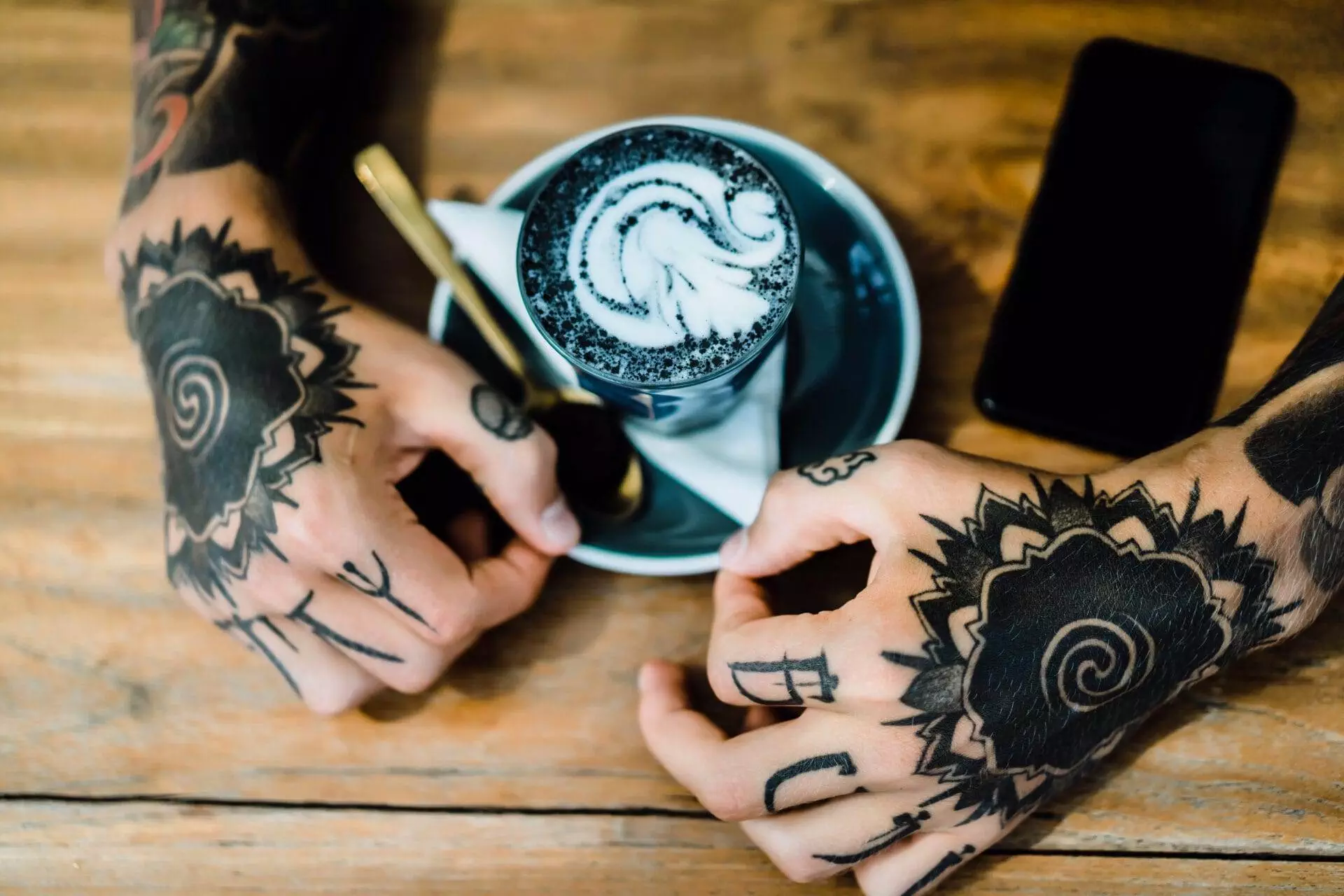 Tattooed hands holding a saucer with a cup of latte art, next to a smartphone on a wooden table.