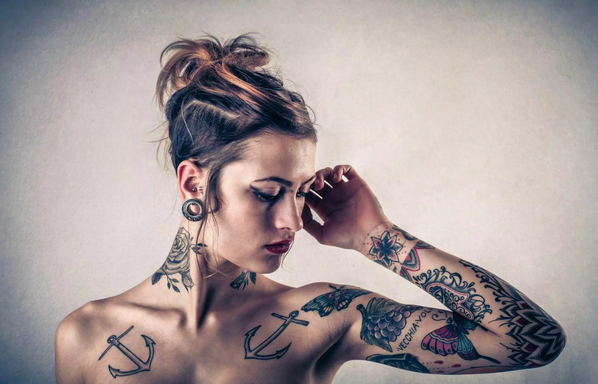 A person with tattoos, including anchors on the shoulders and various designs on the arms, poses with one hand touching their face against a neutral background.