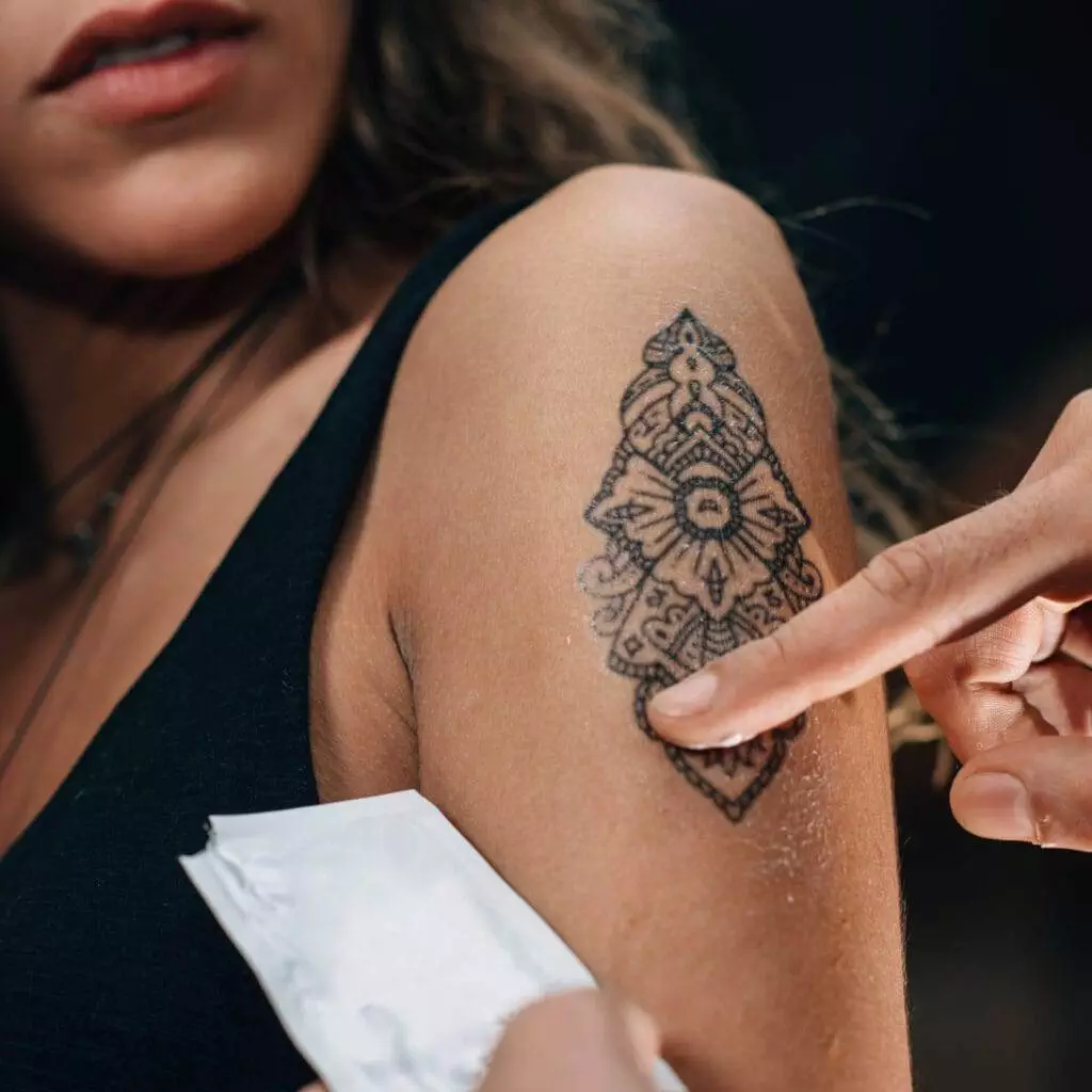 Close-up of a person's arm with a black patterned tattoo. Another hand is pointing at the tattoo while the person wearing a black sleeveless top looks on.