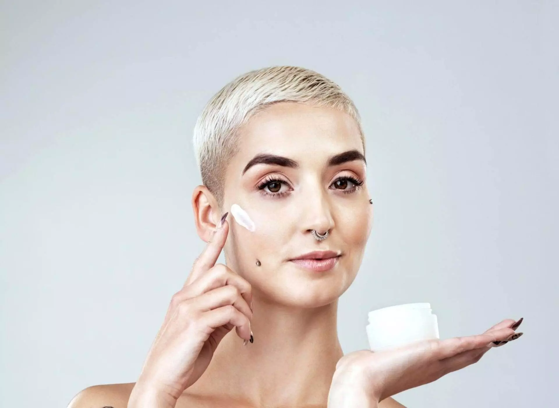 A person with short blonde hair applies a dab of cream to their face, holding a small open jar of the cream in the other hand.