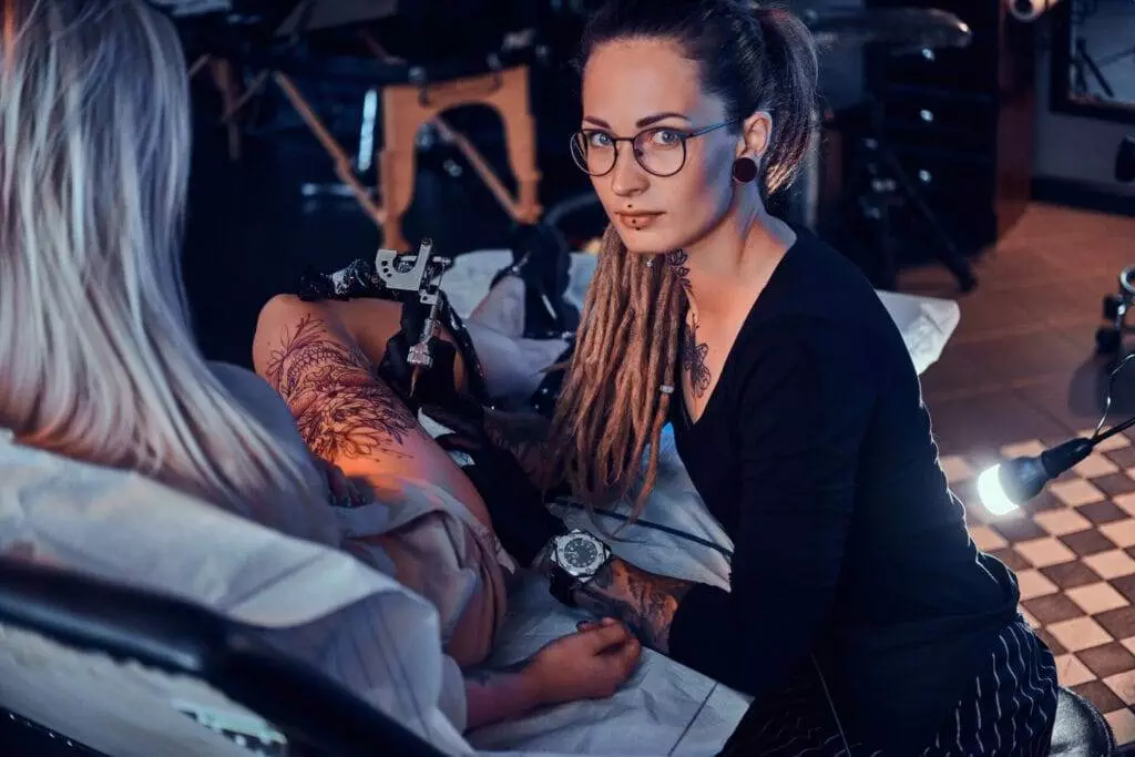 A tattoo artist with glasses and dreadlocks works on a tattoo on a client's thigh in a tattoo studio.