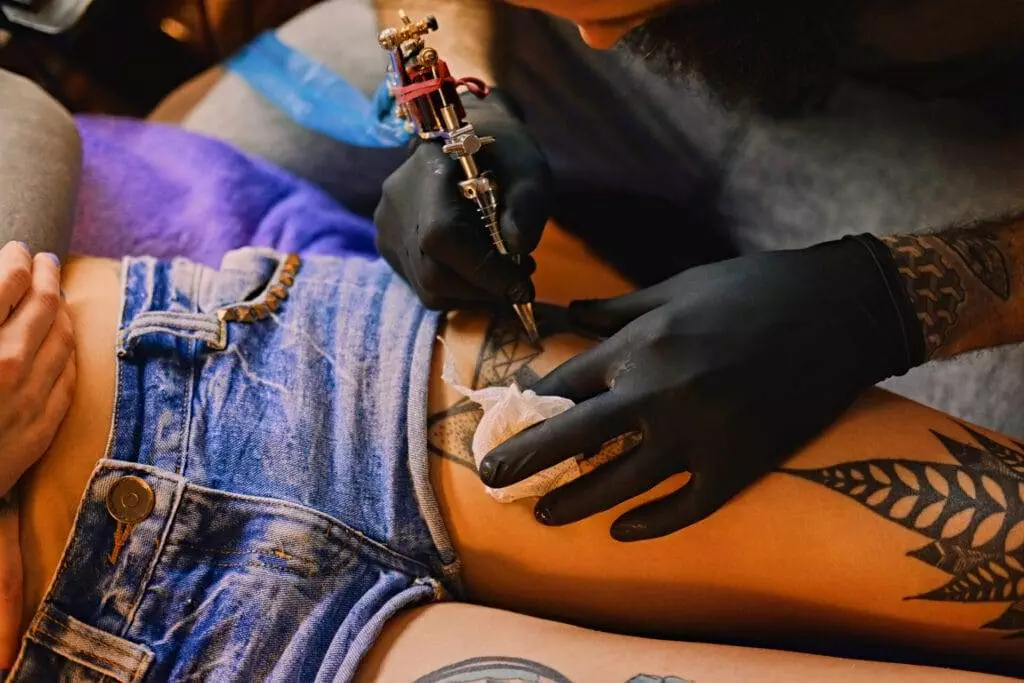 An arm with tattoos is tattooing a design on someone's upper thigh while they lie on a couch, wearing denim shorts.