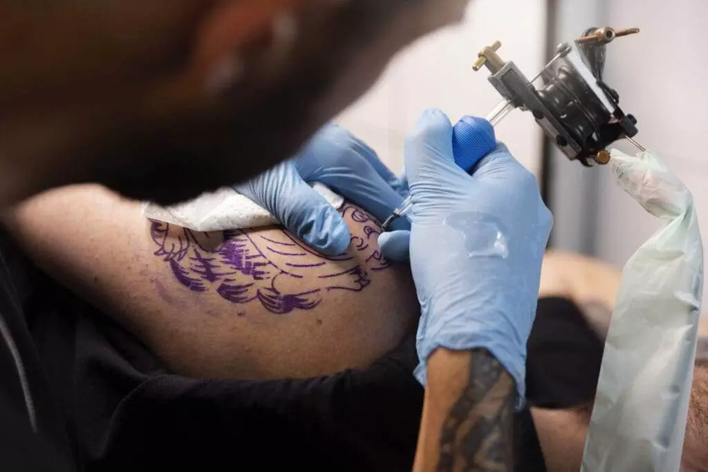 A tattoo artist, wearing blue gloves, uses a tattoo machine to create a detailed design on a person's upper arm.