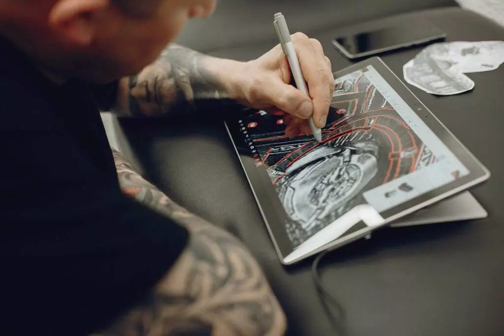 A person with tattoos using a stylus to draw on a tablet, with a phone and paper on the desk beside them.