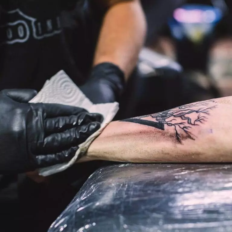 A tattoo artist wearing black gloves wipes a newly inked tree tattoo on a person's forearm with a paper towel.