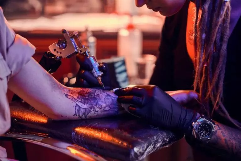 A tattoo artist with dreadlocks and black gloves is working on a client's forearm tattoo in a dimly lit studio.