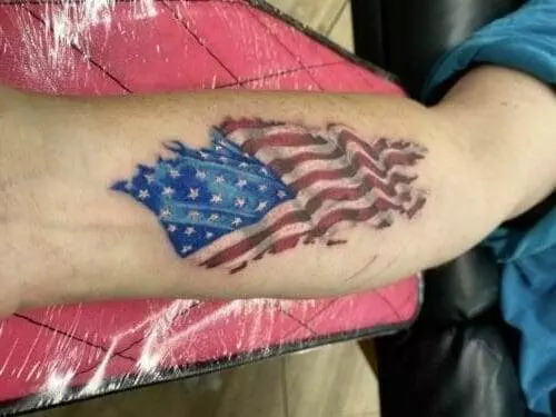 A forearm tattoo depicting a tattered American flag in shades of red, white, and blue, with the arm resting on a red and black background.