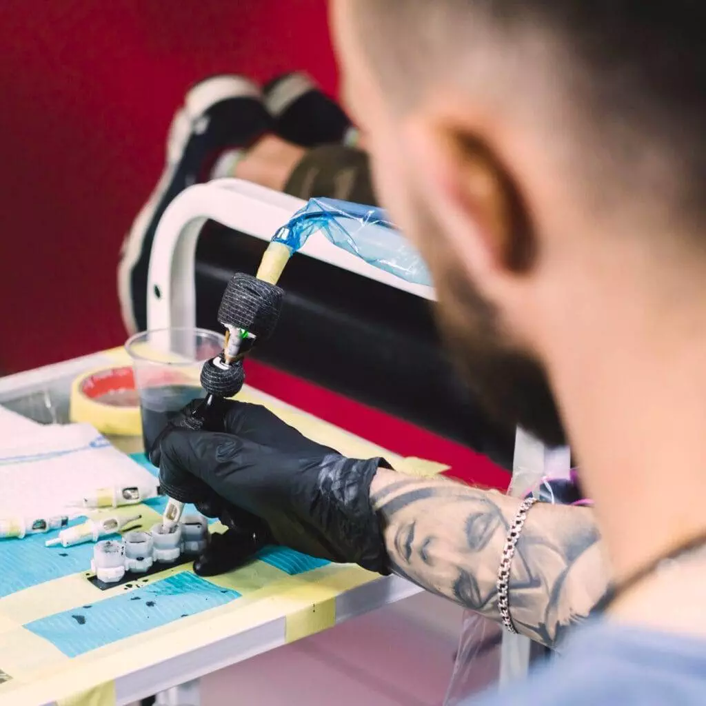 Person with tattoos on their arm and wearing gloves is preparing a tattoo machine beside containers of ink and a cup.