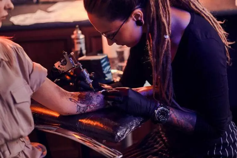 A tattoo artist with long dreadlocks is focused on creating a design on a client's forearm, using a tattoo machine. The client is seated comfortably, their arm resting on a padded surface.