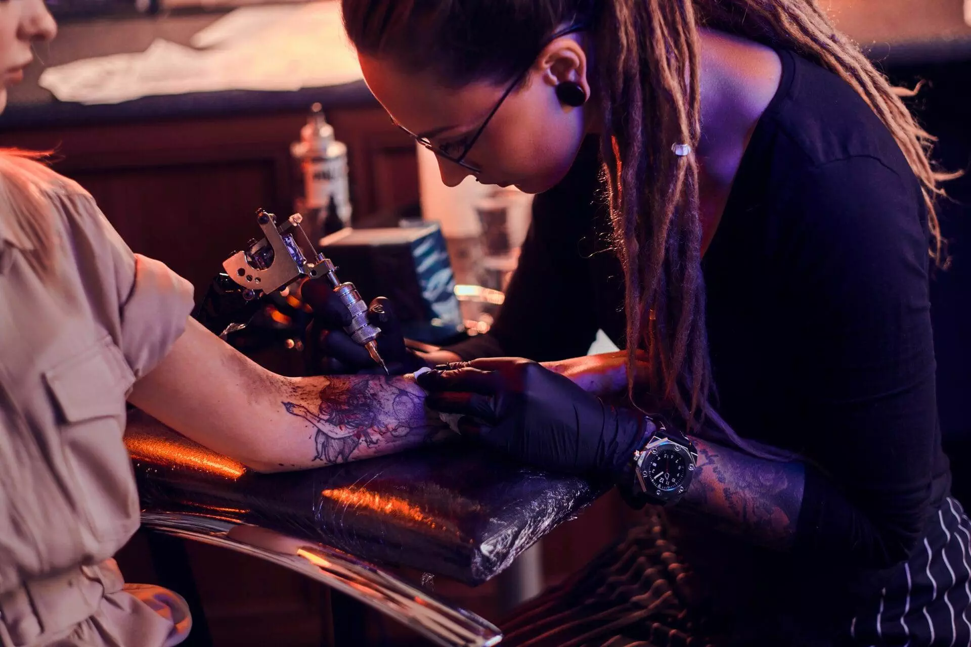 A tattoo artist with long dreadlocks is focused on creating a design on a client's forearm, using a tattoo machine. The client is seated comfortably, their arm resting on a padded surface.