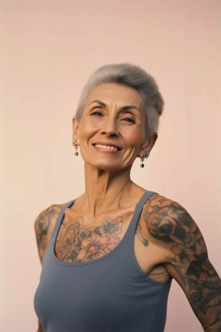 A woman with gray hair and floral tattoos on her arms and chest smiles while wearing a blue tank top.