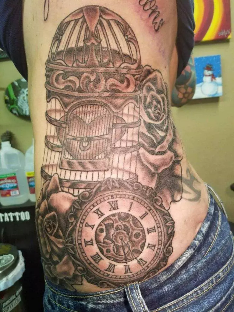 A grayscale tattoo on a person's side featuring an ornate birdcage, roses, and a pocket watch with Roman numerals.